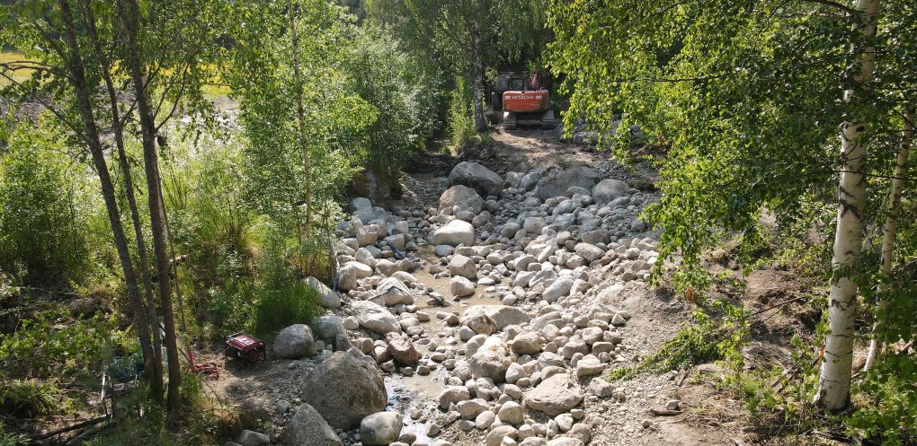 Uutta purouomaa rakennetaan oranssilla kaivinkoneella. Kiviä uoman pohjalla, ympärillä metsää. 