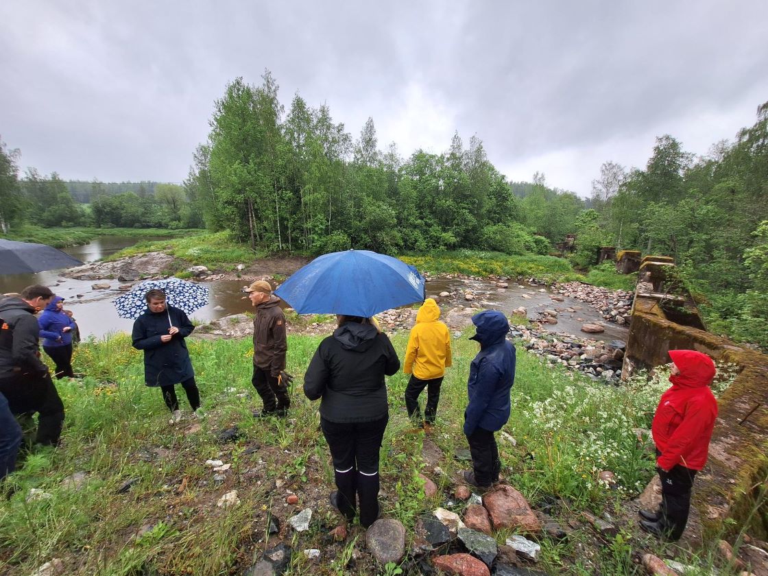 Ihmisiä joen rannassa, jossa taustalla padon rakenteita ja kunnostettua koskenniskaa.