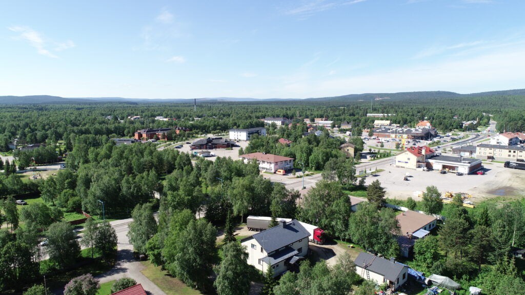 Kuvassa näkyy etualalla taloja puiden seassa. Keskellä kuvaa on katuja ja rakennuksia Ivalon keskustassa. Taustalla näkyy vaaramaisemaa ja sininen taivas.